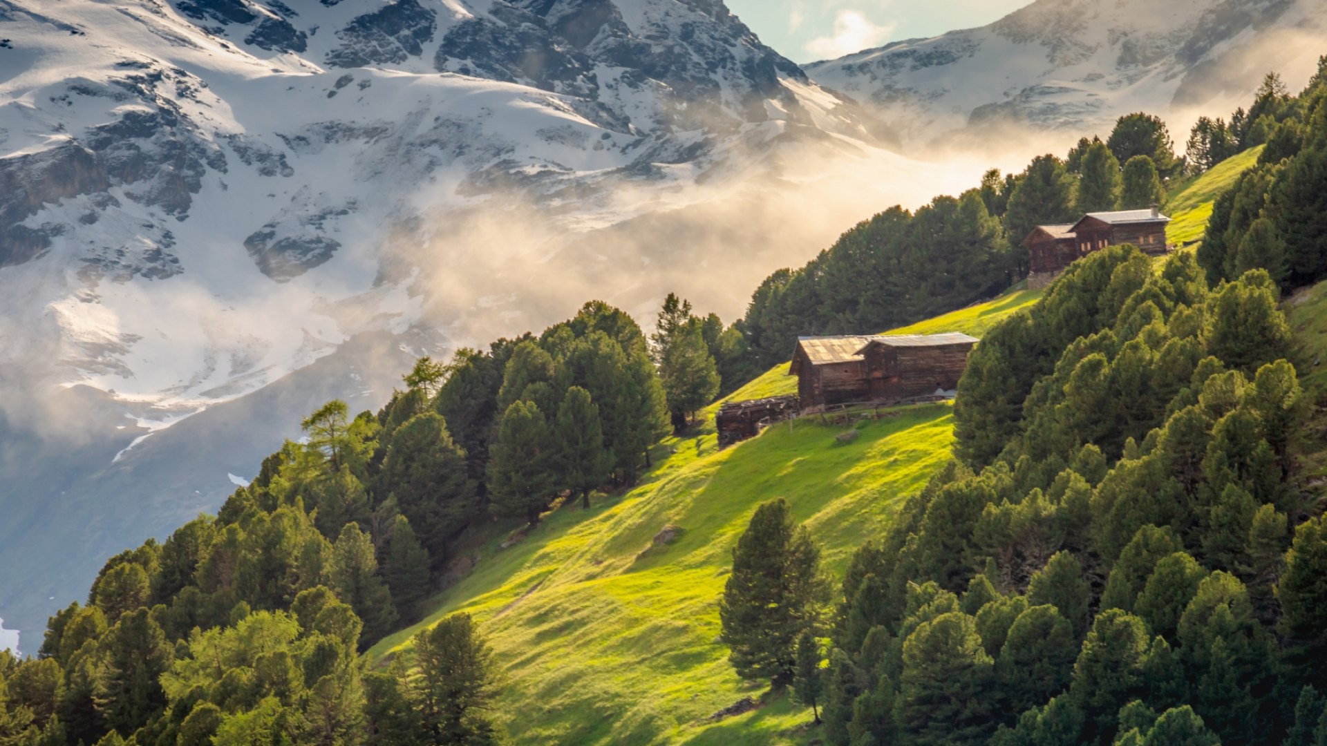 Chalet alpini su un pendio verde, con montagne innevate sullo sfondo.