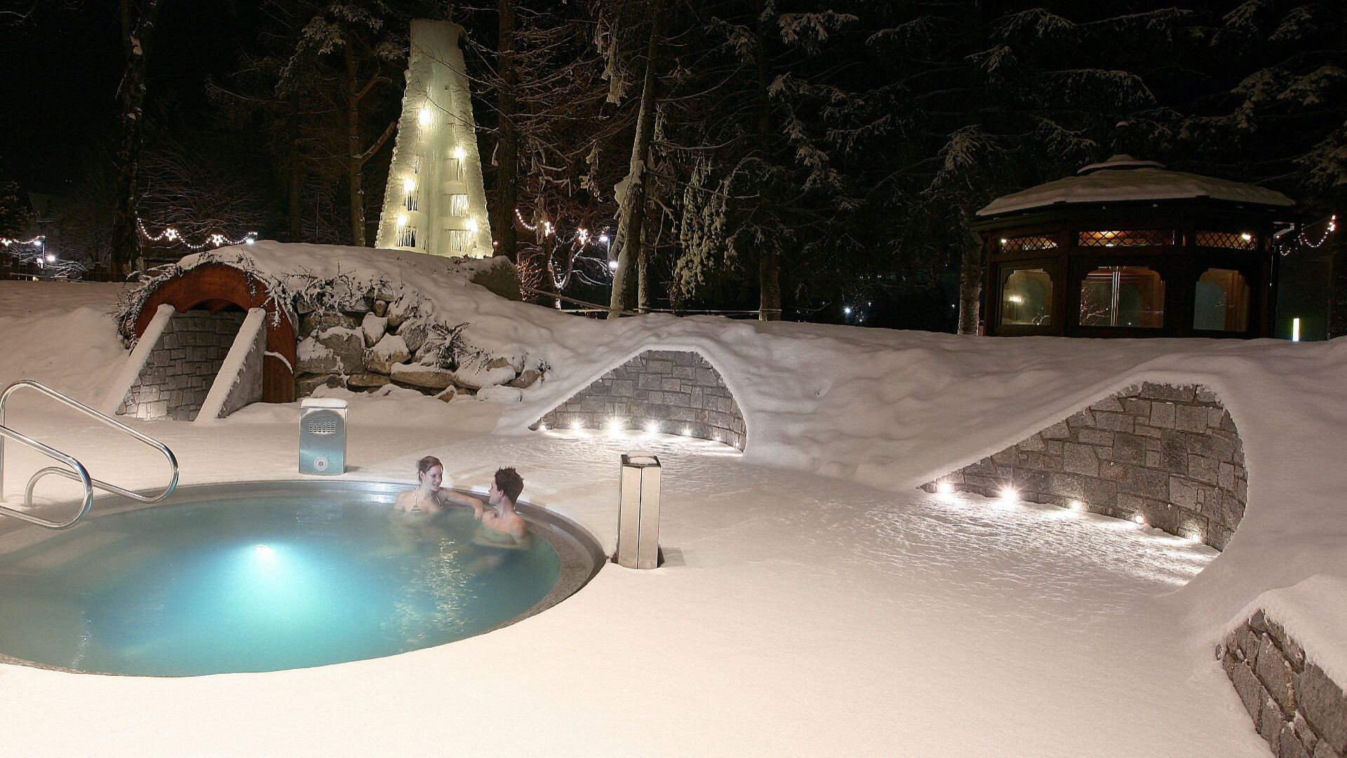 Due persone in una piscina termale all'aperto, circondate da neve e luci.