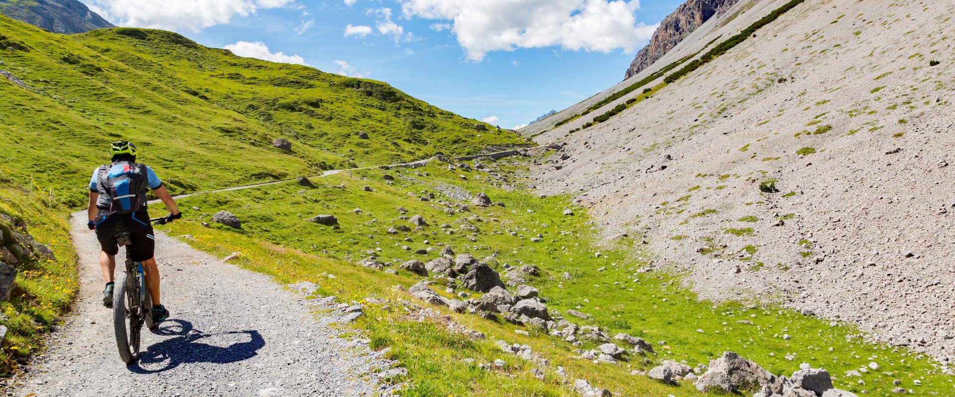 Ciclista su un sentiero di montagna, circondato da prati verdi e rocce.