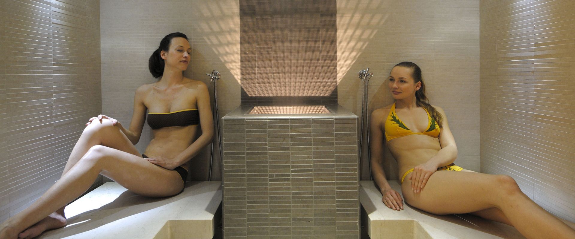 Due donne in bikini si rilassano in una sauna con pareti piastrellate.