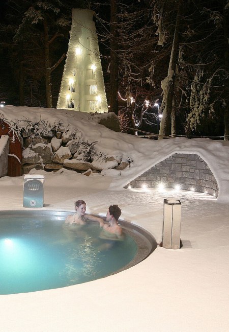 Due persone in una piscina termale all'aperto, circondate da neve e luci.