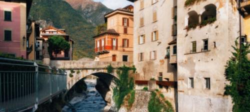 Vecchio ponte in pietra tra edifici storici e fiume, montagna sullo sfondo.