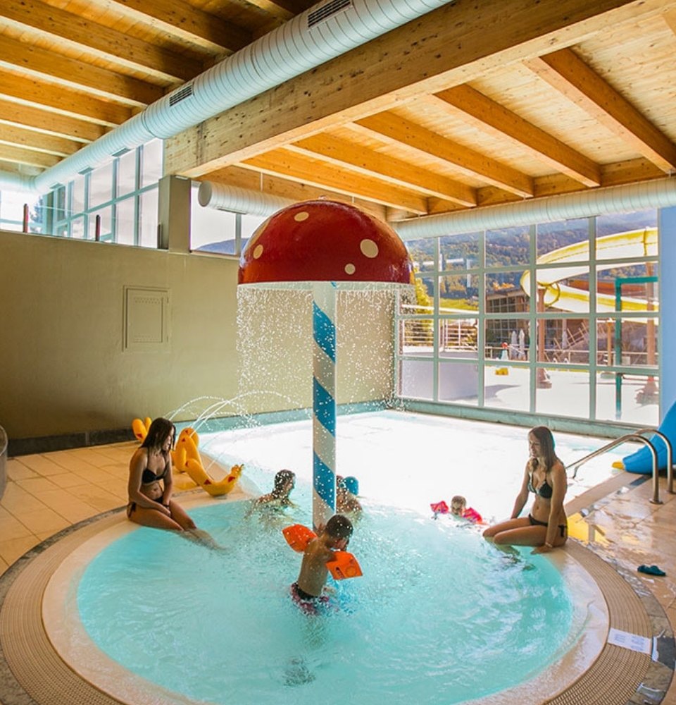 Piscina interna con fungo d'acqua, bambini che giocano e adulti che li sorvegliano.