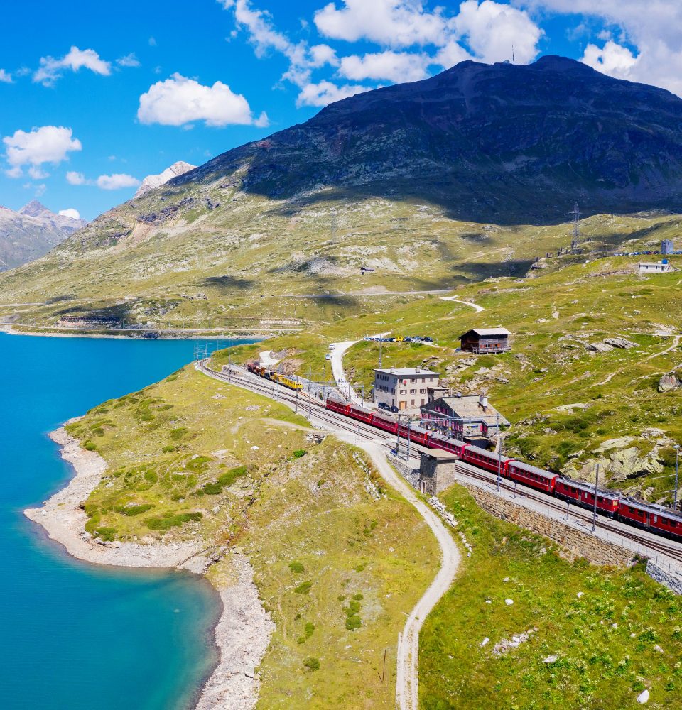 Treno rosso viaggia tra montagne e lago blu sotto un cielo sereno.