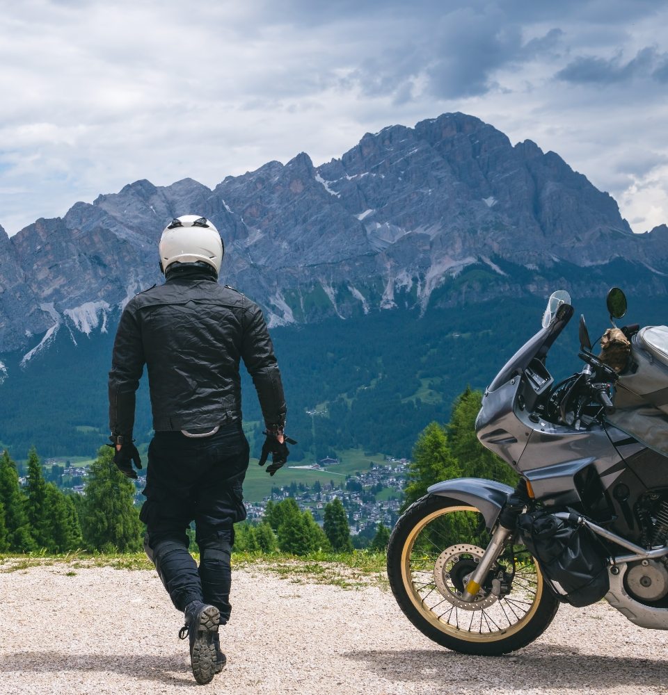 Motociclista con casco osserva montagne, moto parcheggiata su strada sterrata, paesaggio alpino.