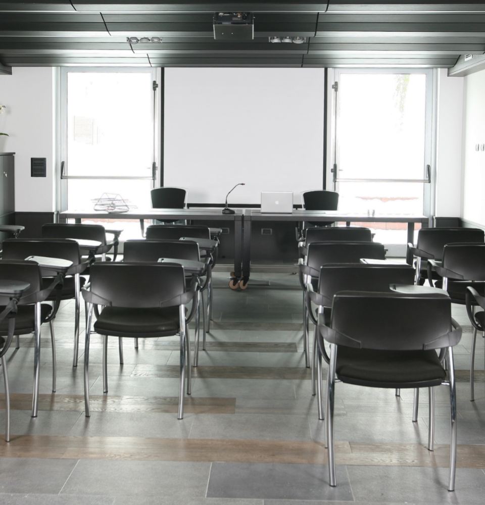 Aula conferenze con sedie nere, tavolo, microfono e laptop, pronta per una presentazione.