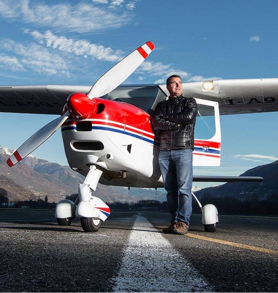 Uomo accanto a un aereo leggero su una pista con montagne sullo sfondo.